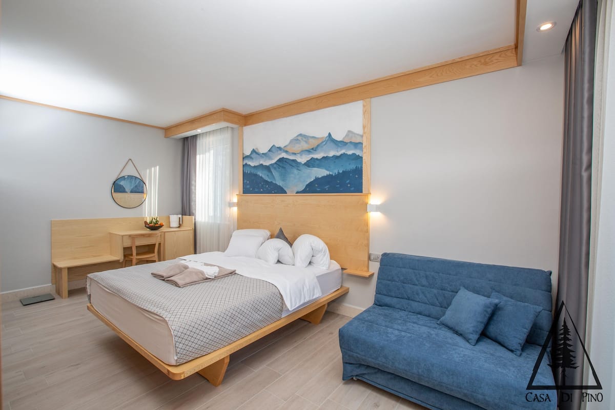 Une chambre moderne avec un lit en bois, un canapé bleu, un bureau et un tableau représentant des montagnes.