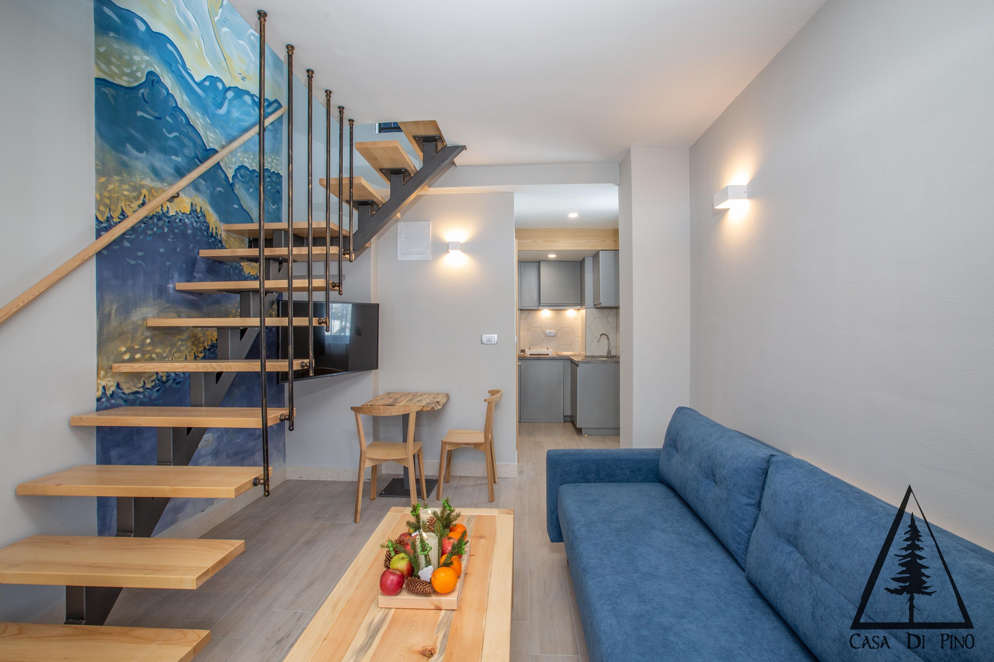 A modern living room with a blue sofa, wooden staircase, and a dining table with fruit.