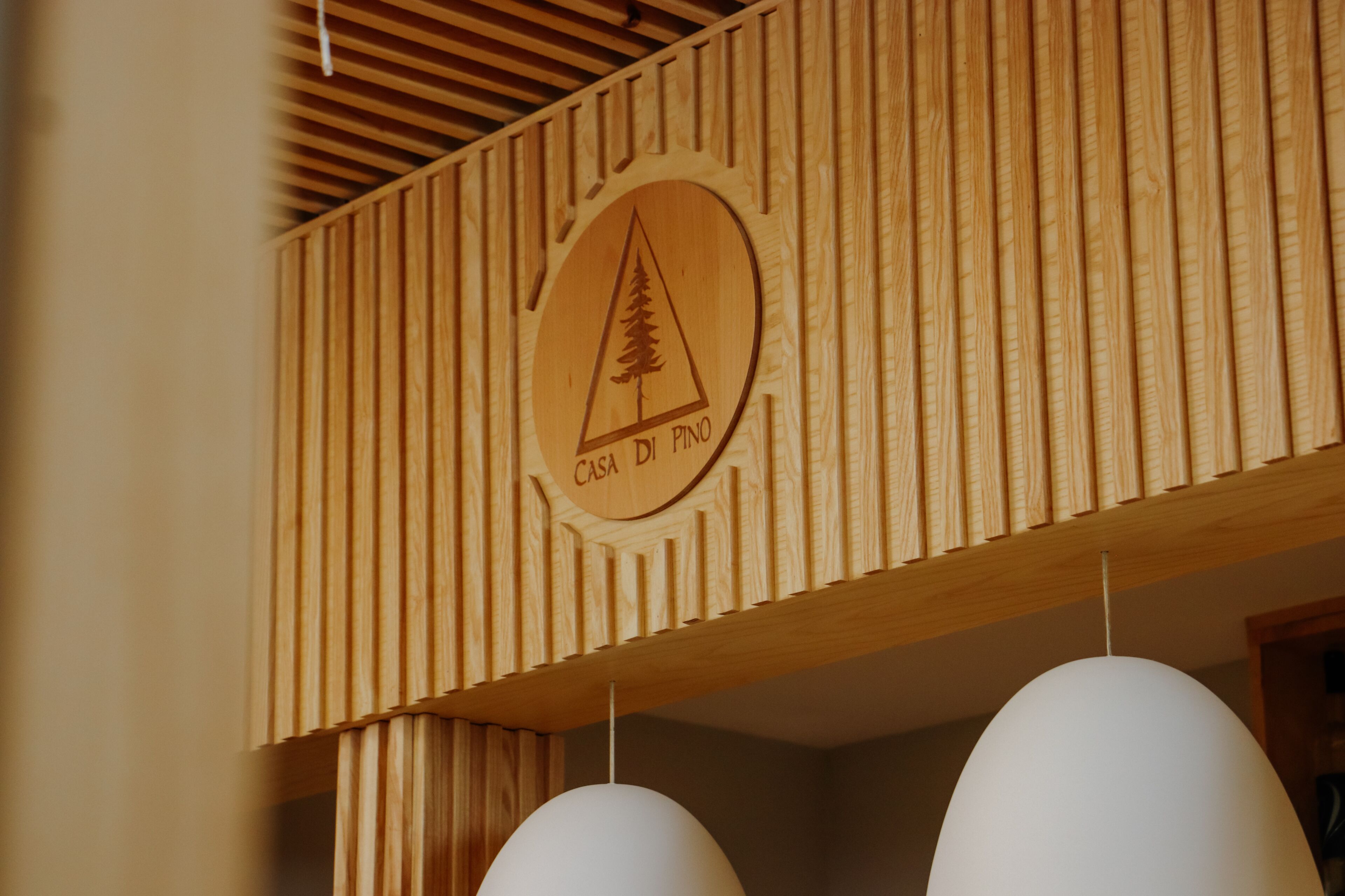 Een houten wand met een rond logo met een pijnboom en de tekst 'Casa Di Pino'.