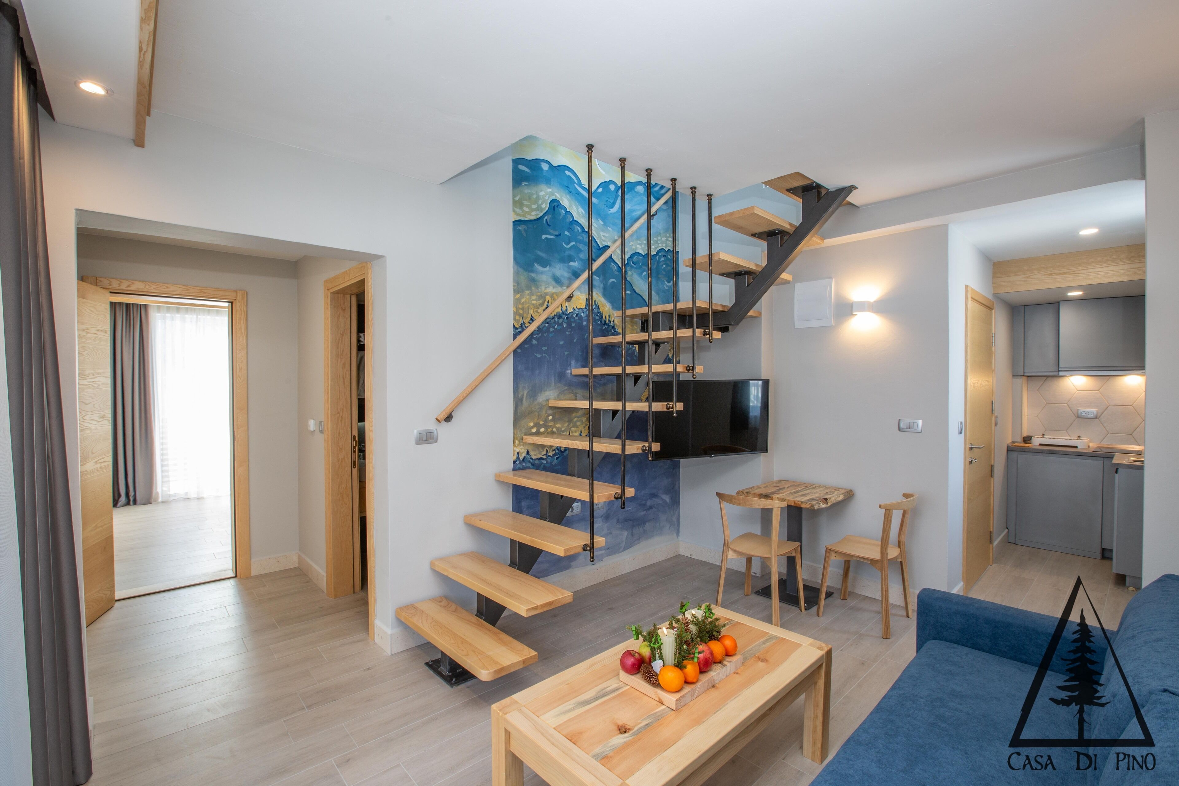 Una sala de estar moderna con una escalera de madera, un comedor con un frutero y una cocina al fondo.