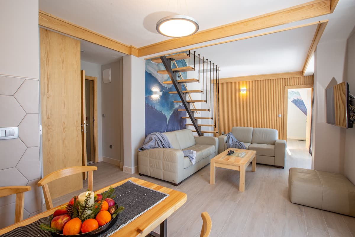Un salon moderne avec un escalier en bois, deux canapés, une table à manger ornée de fruits et un tableau représentant un paysage de montagne.