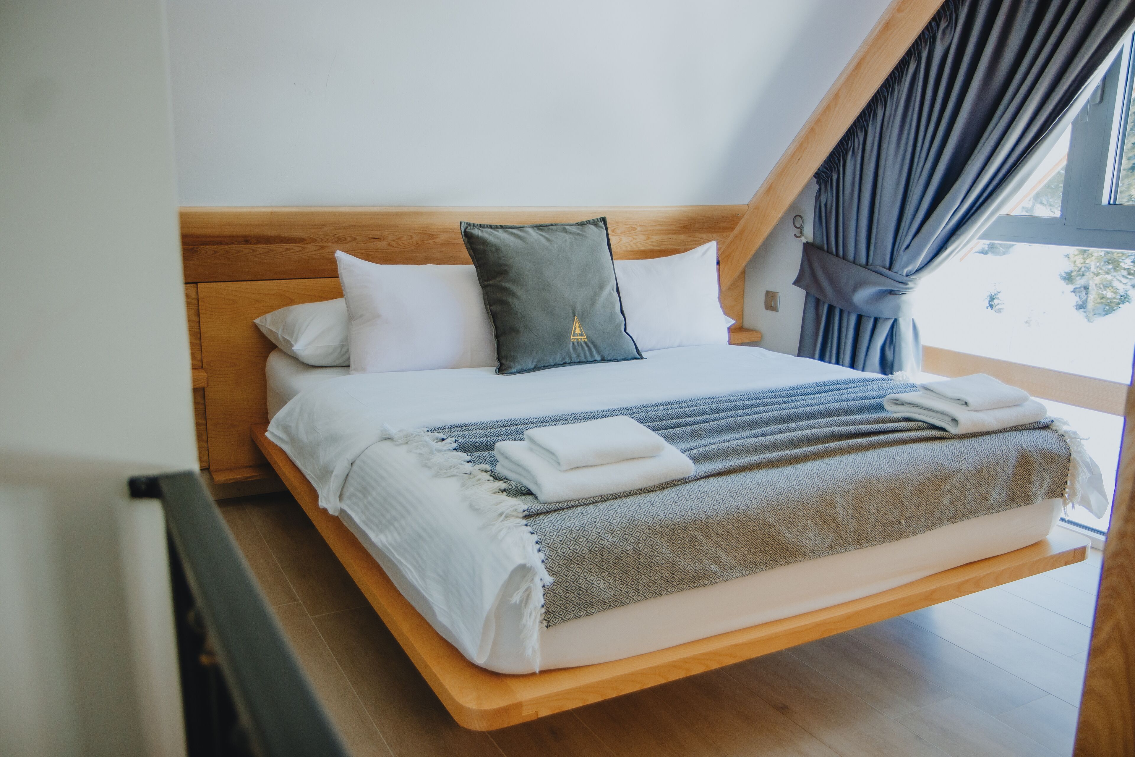 A bed with a wooden headboard, white and gray bedding, and a view of the outdoors through a window with curtains.