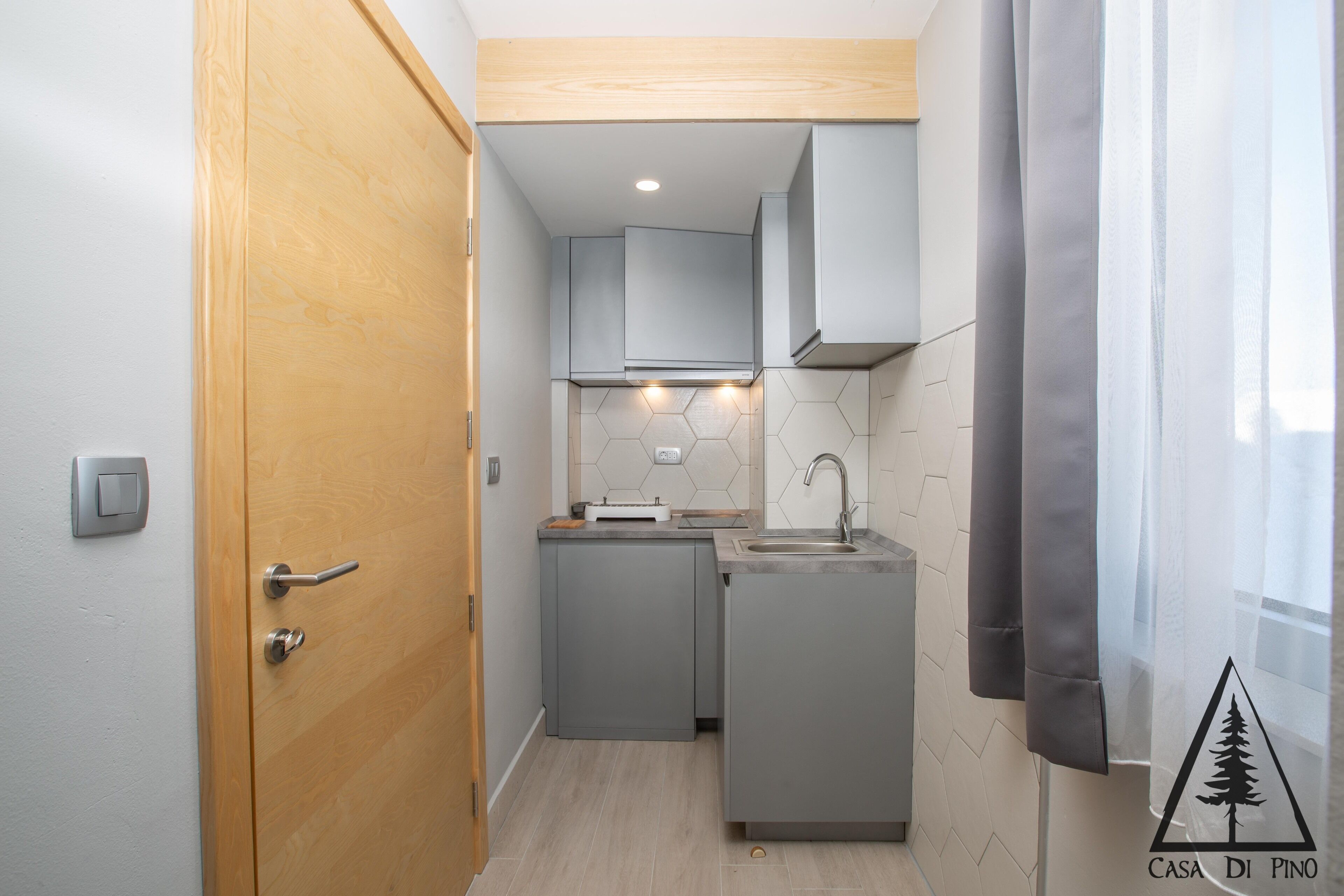 A compact kitchen with grey cabinets, a wooden door, and a window with a curtain.