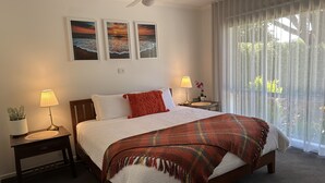Un lit bien fait, recouvert d’une couette blanche et d’un plaid rouge, deux lampes de chevet, une petite plante en pot et trois photos encadrées représentant des couchers de soleil, accrochées au mur.