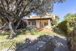 Ein Haus im mediterranen Stil mit einem Kiesweg, der zum Eingang führt, umgeben von üppiger Vegetation und einem großen Baum.