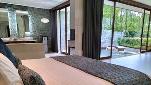 A modern bedroom with a large bed, a bathroom with a sink and mirror, and a view of a garden through large windows. - Rama Gaia Villa (Ubud)