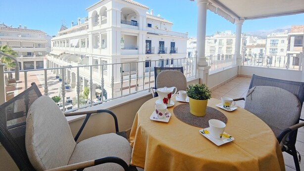 A balcony with a table set for two, chairs, a potted plant, and a view of surrounding buildings.
