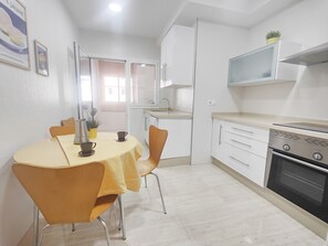 Una cocina compacta con armarios blancos, estufa y microondas. Hay una mesa de comedor con sillas y mantel amarillo.