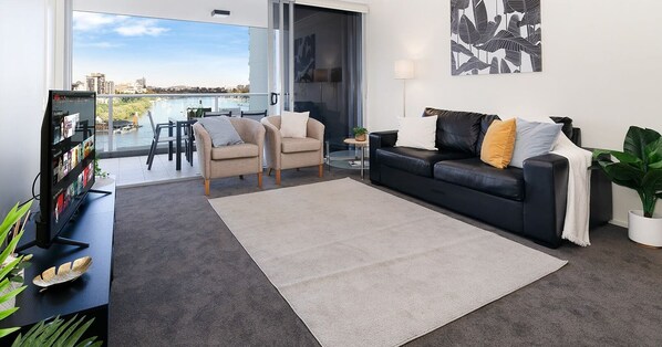 Ein modernes Wohnzimmer mit großem Fernseher, zwei Sesseln, einem schwarzen Ledersofa, einem Essbereich mit Tisch und Stühlen und Blick auf ein Gewässer durch große Fenster.