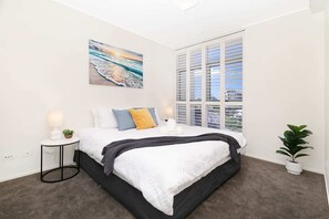 A bedroom with a bed, a nightstand, a lamp, a plant, and a painting of a beach scene. - Executive Family Suite View Parking WiFi (Brisbane)