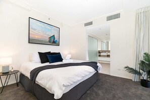 Ein Hotelzimmer mit einem großen Bett, einem Nachttisch mit Lampe, einem Wandbild mit Booten und einem durchgegangenen Tür sichtbaren Badezimmer.