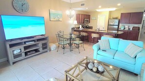 A living room with a television, a dining area with chairs and a table, a kitchen with cabinets and appliances, and a light blue sofa. - Saltwater Retreat, 2nd Floor, Spectacular Pool Area, Hot Tub, Free Beach Service (Miramar Beach)