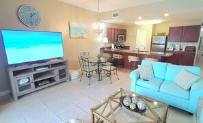 A living room with a television, a dining area with chairs and a table, a kitchen with cabinets and appliances, and a light blue sofa.