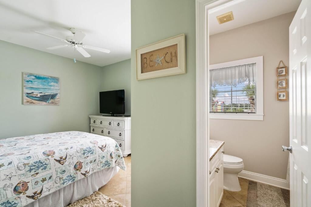 A bedroom with a bed, a television on a dresser, a painting, and a bathroom with a toilet and window.