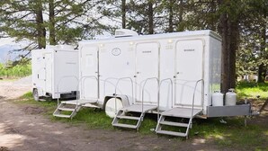 Una fila de camperas blancas equipadas con duchas, ubicadas en un camino de tierra con árboles al fondo.