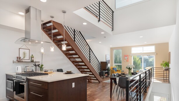 A modern kitchen with a central island, wooden flooring, and a staircase with a black railing.