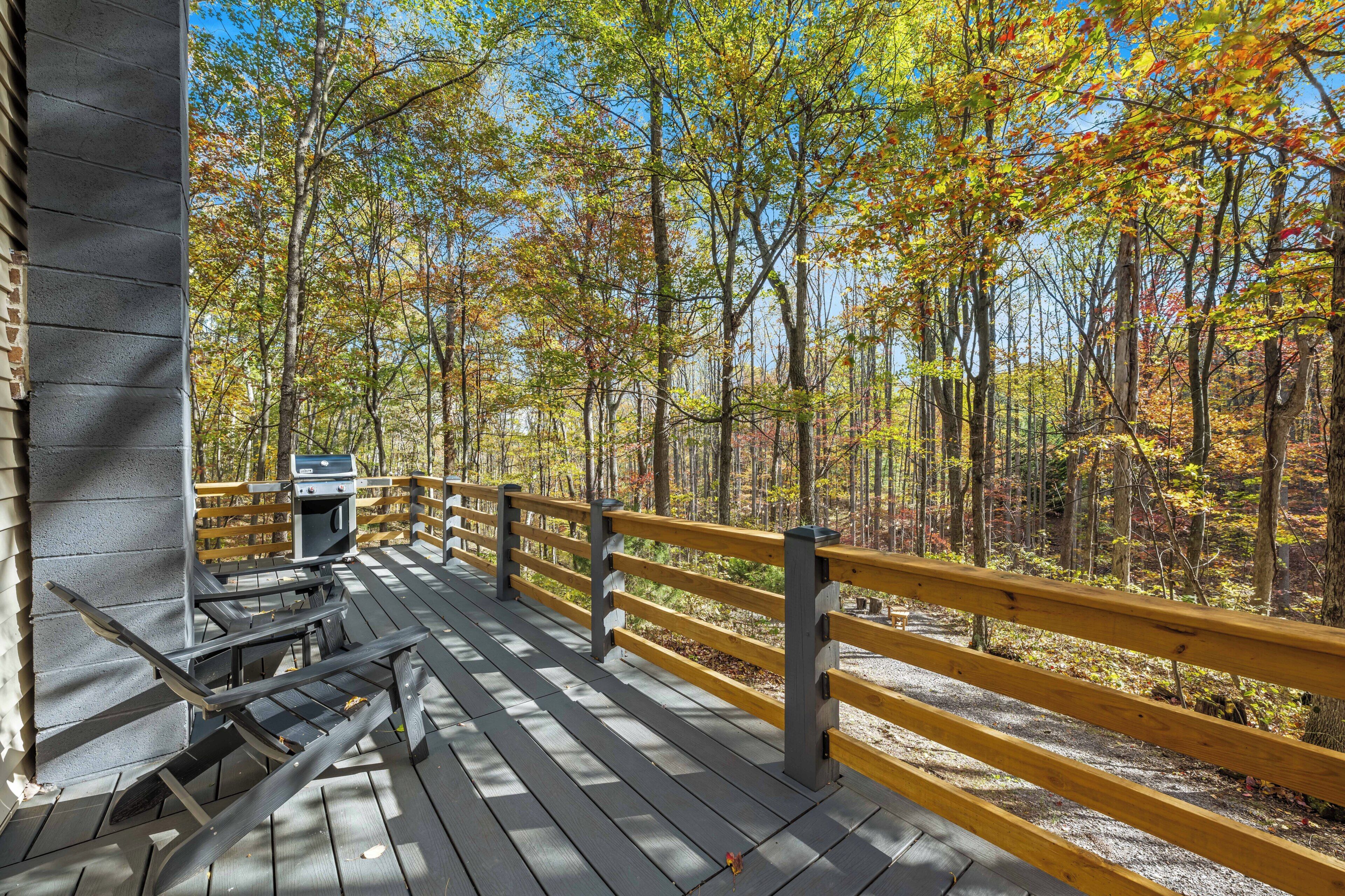 A wooden deck with a grill and picnic table, surrounded by a forest with autumn foliage.