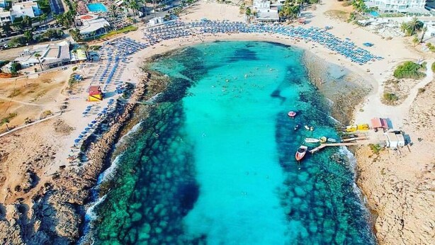 An aerial view of a coastal area with a clear turquoise sea, a sandy beach, and several buildings, including hotels and resorts. - Nissi Avenue Apartment in Ayia Napa (Ayia Napa)