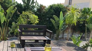 A wooden deck with two wooden benches and a table, surrounded by tropical plants. - Nissi Avenue Apartment in Ayia Napa (Ayia Napa)