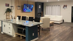 A modern kitchen with a central island, dining area with chairs, a white sofa, and a flat-screen TV mounted on the wall. - CG Suites (Columbus)