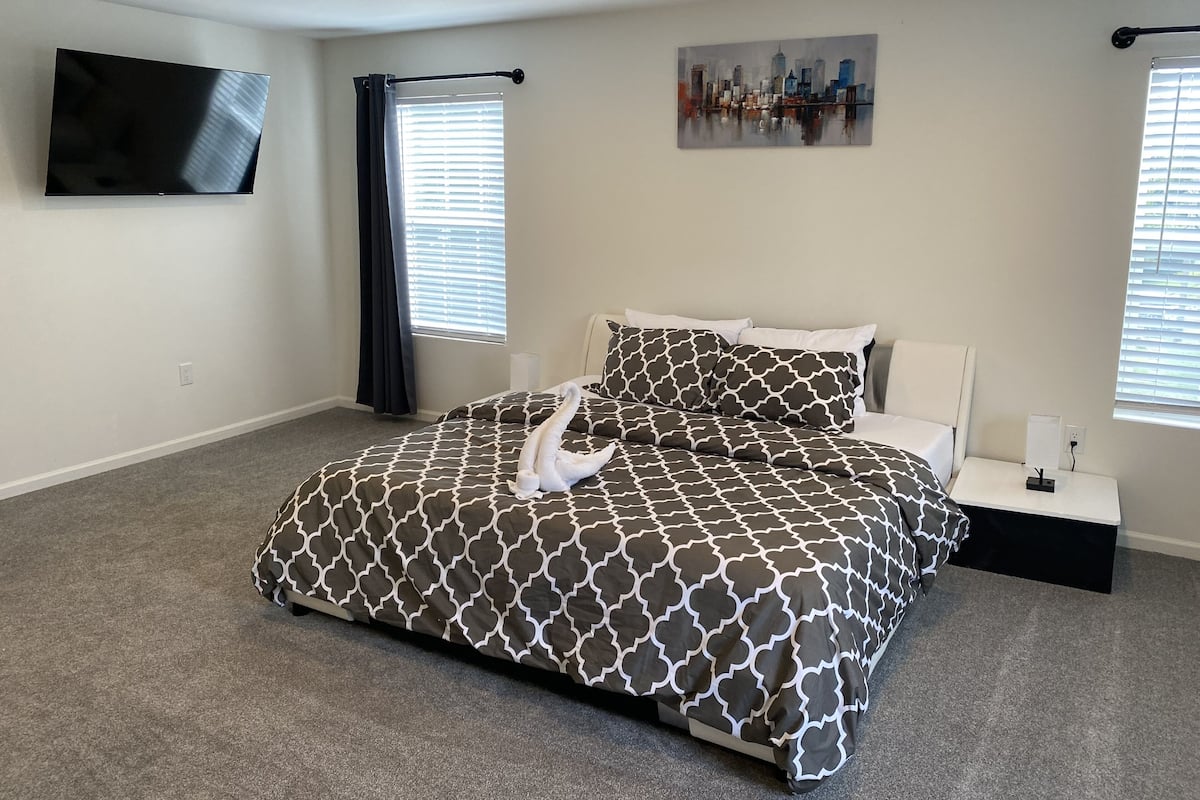 A bedroom with a bed, a flat-screen TV mounted on the wall, a painting, and a window with blinds.