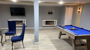 A room with a pool table, two blue chairs, a television, and a fireplace. - CG Suites (Columbus)