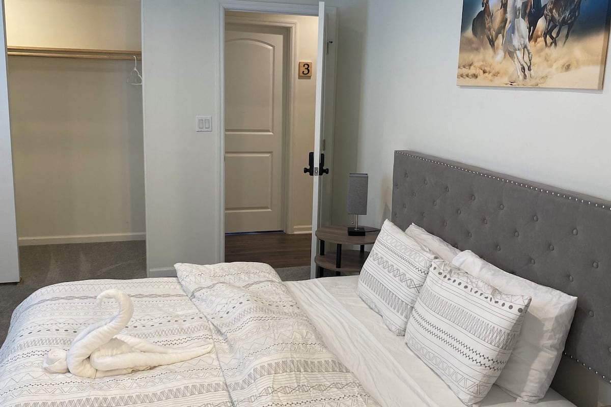 A bedroom with a large bed, a grey upholstered headboard, and a painting of horses on the wall.