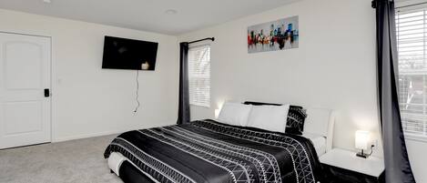 A bedroom with a bed, a TV mounted on the wall, a window with curtains, and a painting.