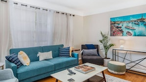 A living room with a teal sofa, a grey armchair, a coffee table, and a painting of a harbor scene. - The Park & River Chalet - Duplex 2 units (Detroit)