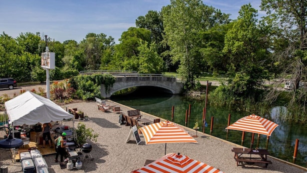 A riverside patio with picnic tables, striped umbrellas, and a covered area with a grill. - The Park & River Chalet - Duplex 2 units (Detroit)