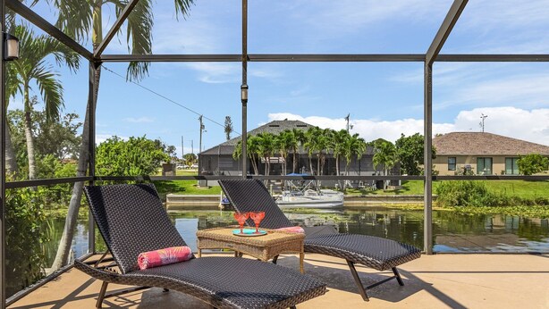 A glass-enclosed lounge area with wicker chairs and a small table, overlooking a body of water and a building with a sloped roof.
