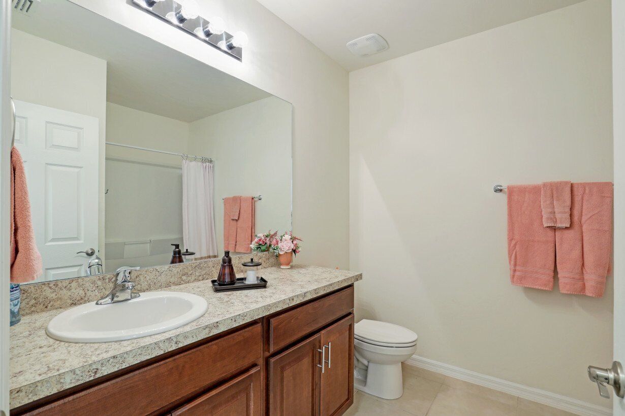 A bathroom with a white sink, a large mirror, and a toilet. There are pink towels hanging on the wall and a shower curtain visible in the background.