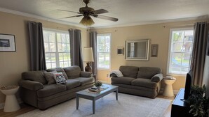 A living room with two sofas, a coffee table, a ceiling fan, and large windows. - Come stay at Suzie's House!<br>Spacious Family-Friendly 4-Bedroom Home (Fort Benton)