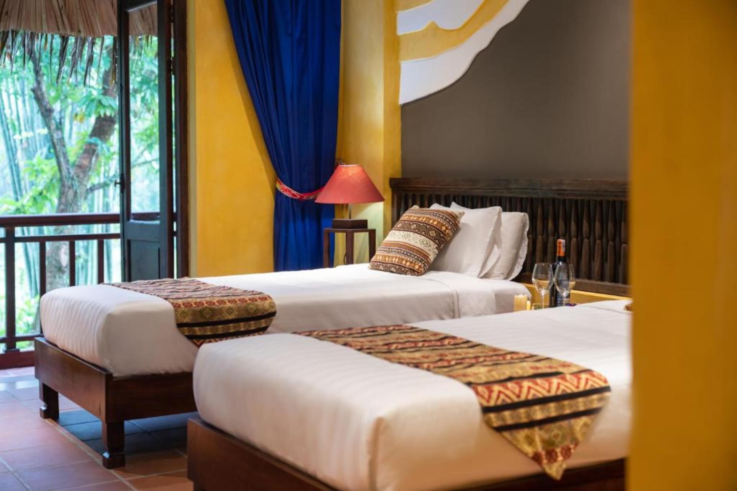Habitación de hotel con dos camas, una mesita de noche con lámpara y vistas a zonas verdes por la ventana.