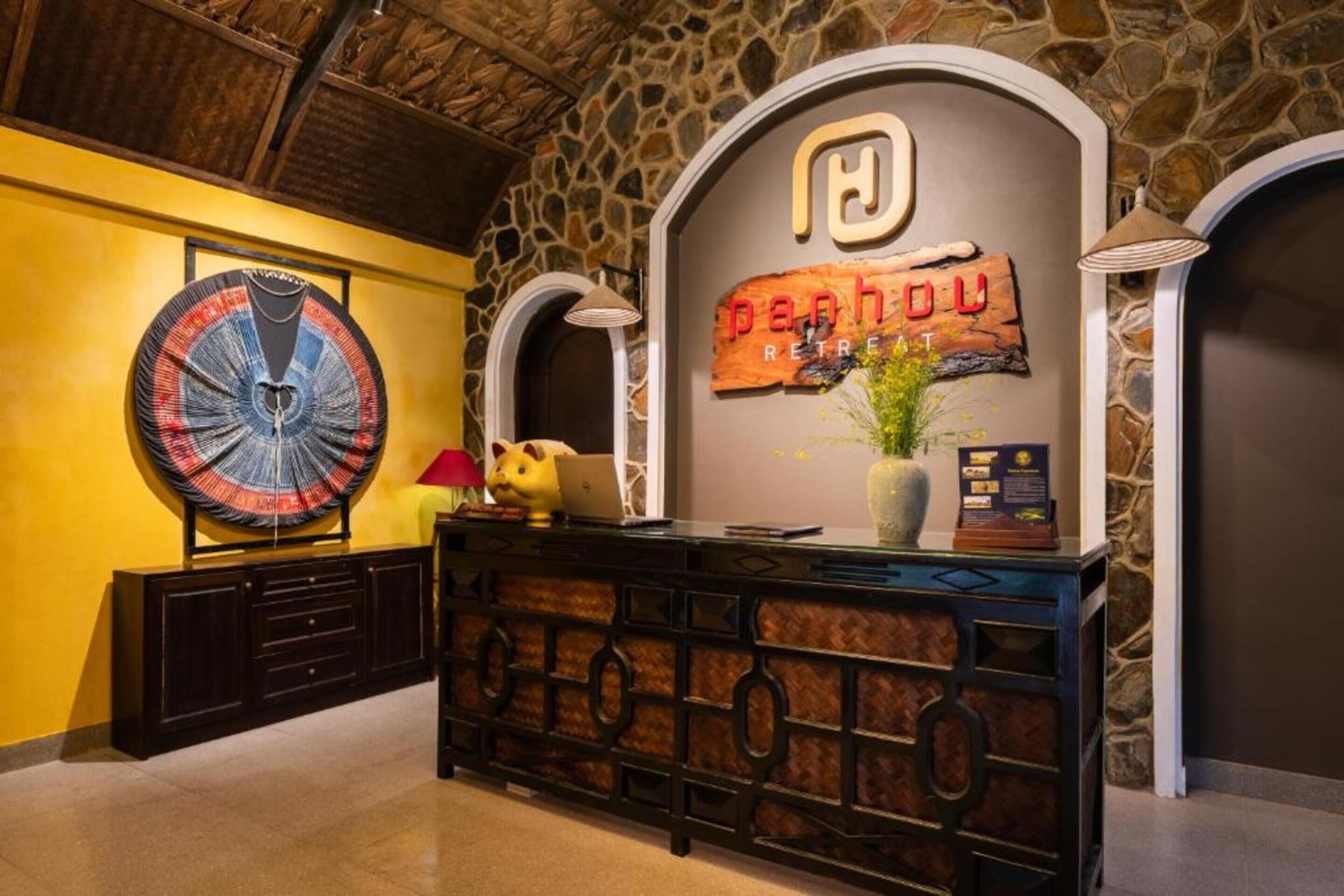 Un área de recepción con pared de piedra, un cuadro abstracto de gran tamaño, un letrero de madera que dice 'panh ou RETRE' y un escritorio con una computadora portátil y un peluche.