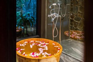 A wooden tub filled with water and rose petals, a shower with a stone wall, and a view of a waterfall through a window.