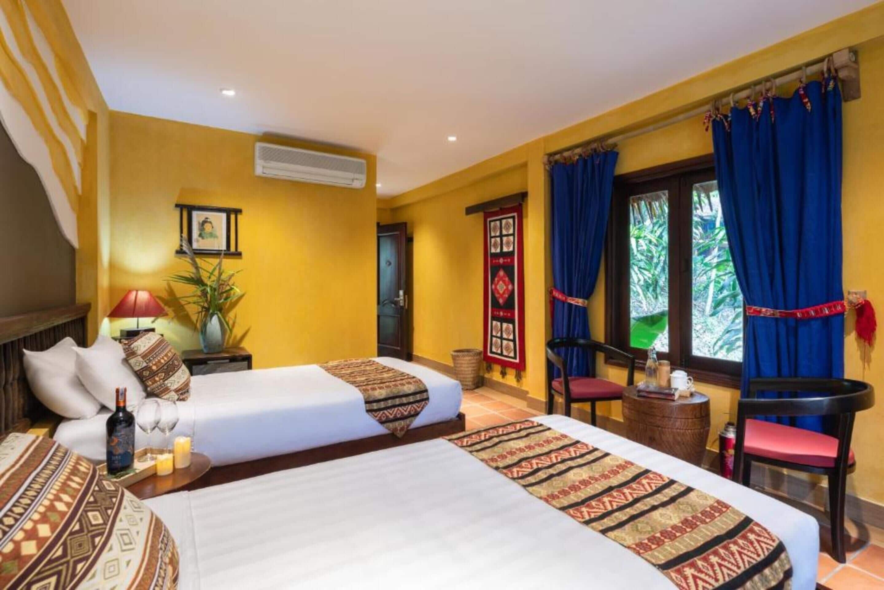 A hotel room with two beds, a small table with a lamp, a chair, and a window with curtains.