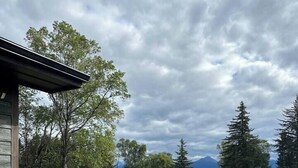 Ein rustikales Blockhaus mit Holzterrasse und Kübelpflanzen, umgeben von einem Schotterweg und einer Rasenfläche mit Bäumen und Bergen im Hintergrund.