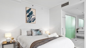 Ein modernes Schlafzimmer mit Bett, Nachttischen, einem Bild und einem durchgehenden Türrahmen einsehbaren Badezimmer.