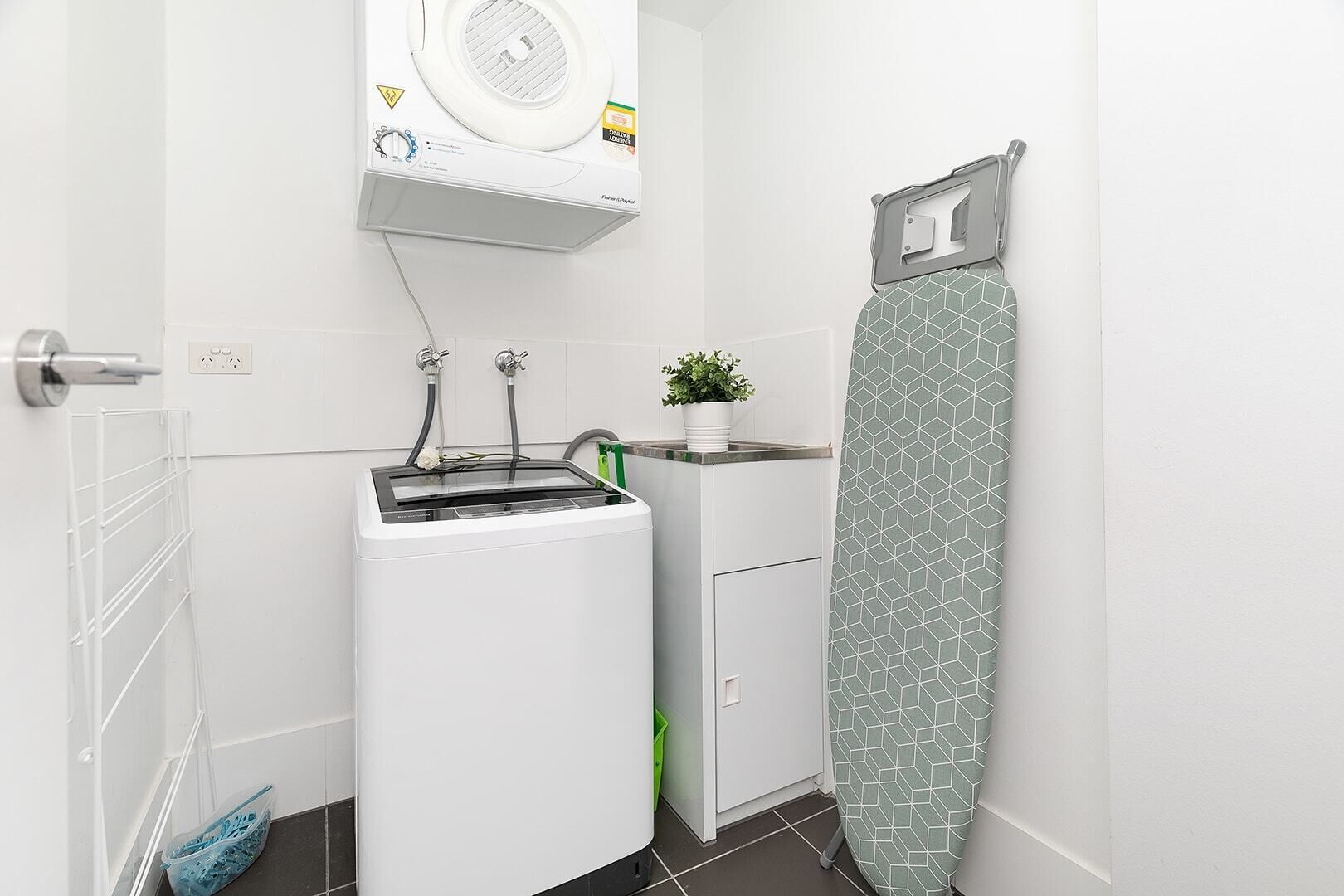 A compact laundry room with a washing machine, a drying rack, and a green plant.