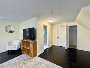 Une pièce avec un téléviseur à écran plat posé sur un meuble en bois, un fauteuil blanc, un tapis à motifs et une porte blanche donnant sur une autre pièce.