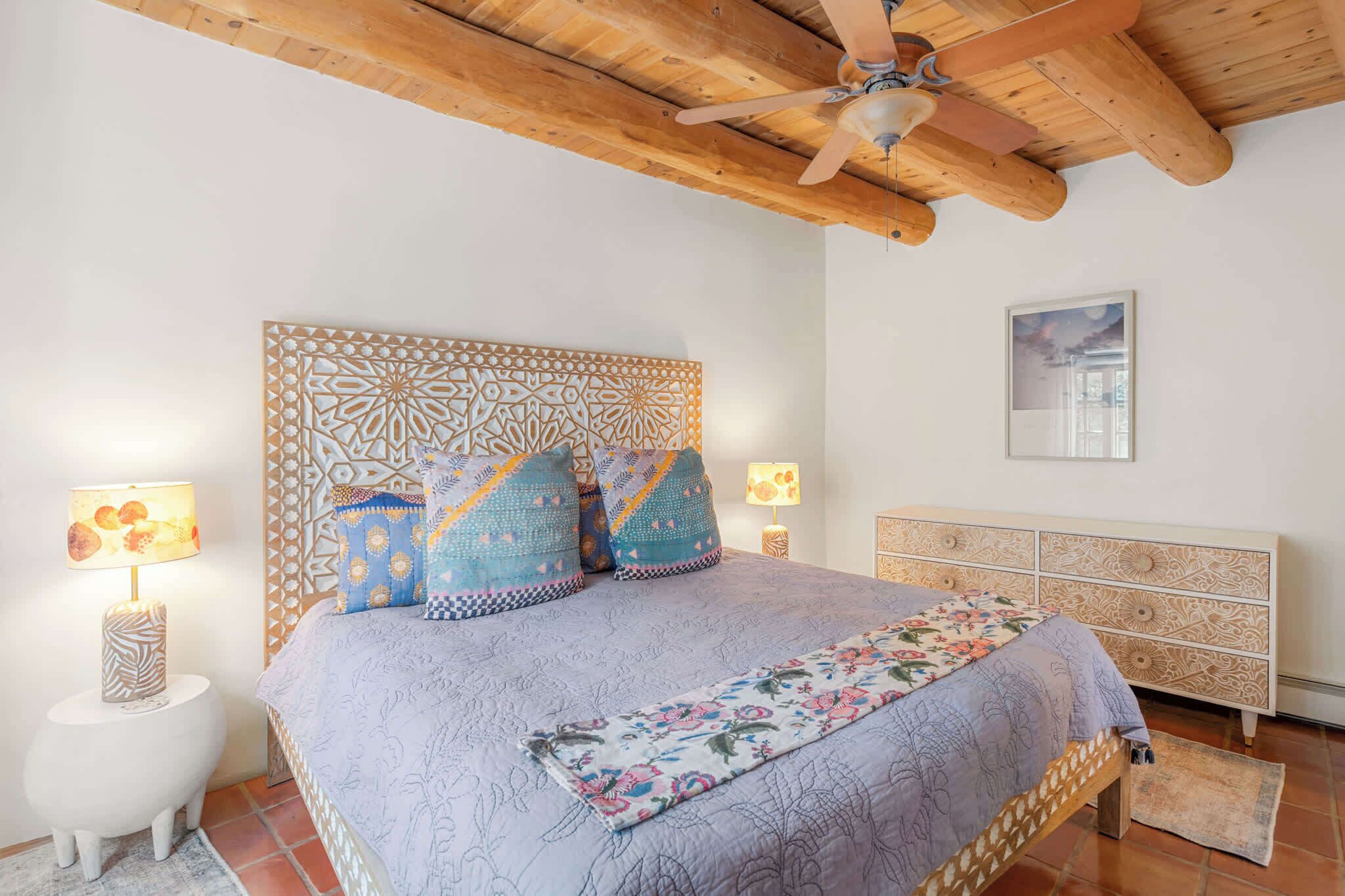 A bedroom with a patterned headboard, a bed with floral bedding, a wooden ceiling fan, and a nightstand with a lamp.
