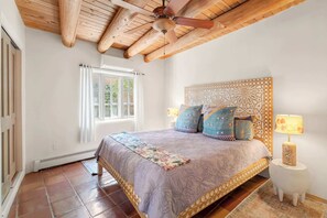 A bedroom with a wooden ceiling, a bed with patterned bedding, a nightstand with a lamp, and a window with curtains. - Amazing Railyard Location, Kiva Fireplace (Santa Fe)