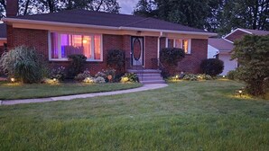 A two-story brick house with a front porch, a well-maintained lawn, and a small garden with various plants and shrubs.