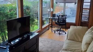 A room with a TV on a stand, a beige sofa, and a desk with a chair. Large windows offer a view of greenery and a building outside.