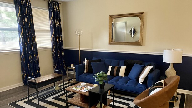 A living room with a blue sofa, a wooden coffee table, a brown armchair, a black and white striped rug, and a floor lamp. - Marines peaceful retreat, The Spiral @ AbbeyAcres Near Beach with Washer & Dryer (Holly Ridge)