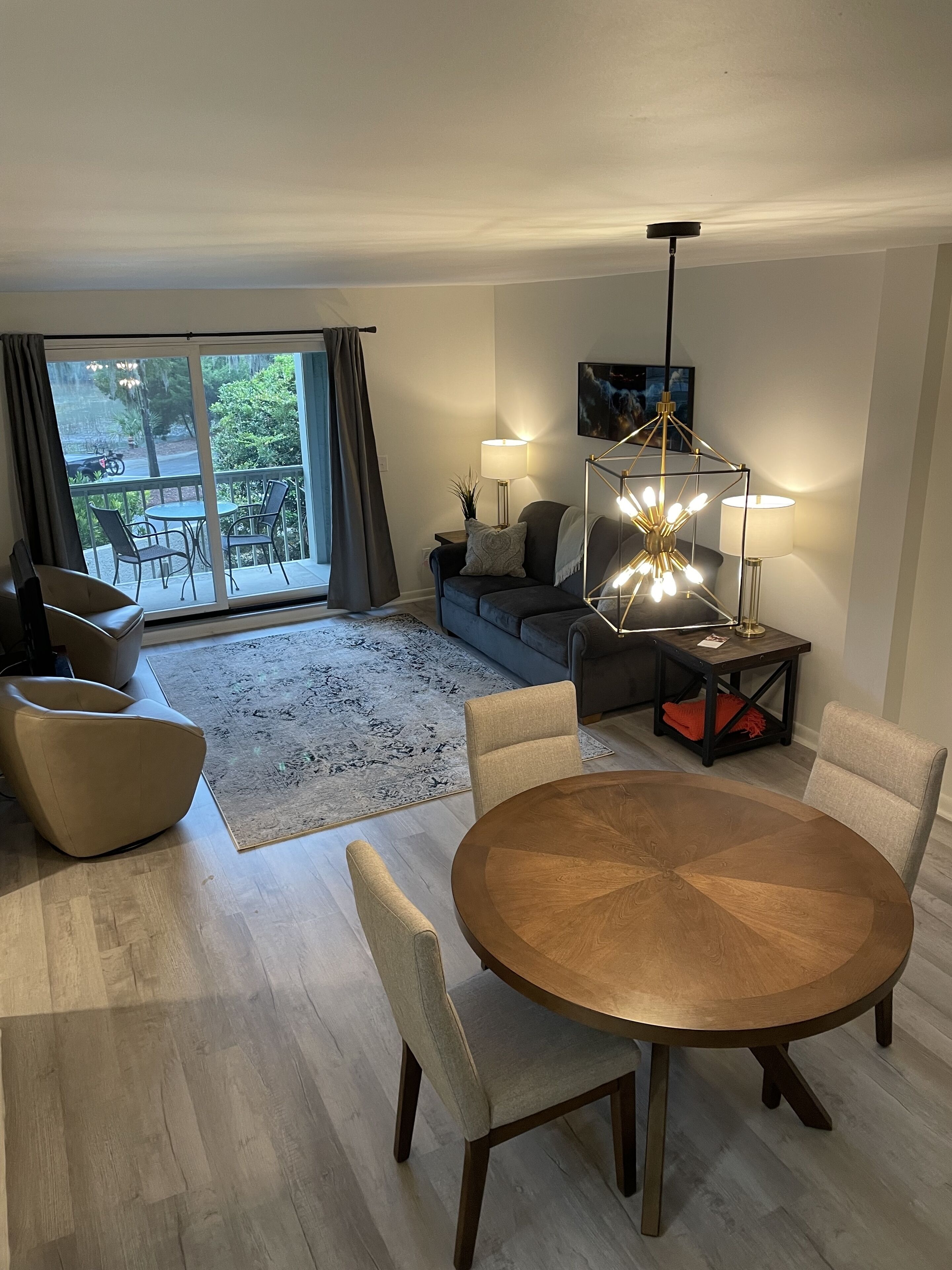 Ein modernes Wohnzimmer mit einem runden Holzesstisch, zwei beigen Sesseln, einem grauen Sofa und einem schwarzen Couchtisch. Es gibt ein großes Fenster mit Vorhängen, eine Stehlampe und ein Gemälde an der Wand.