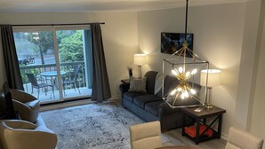 A modern living room with a round wooden dining table, two beige armchairs, a grey sofa, and a black coffee table. There is a large window with curtains, a floor lamp, and a painting on the wall. - Spacious Condo in the heart of Harbor Town! (Hilton Head Island, USA)