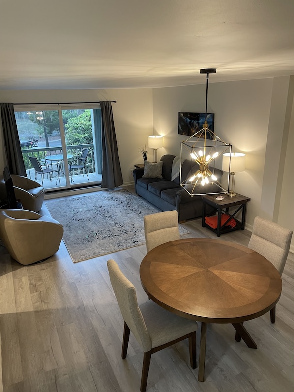 A modern living room with a round wooden dining table, two beige armchairs, a grey sofa, and a black coffee table. There is a large window with curtains, a floor lamp, and a painting on the wall.