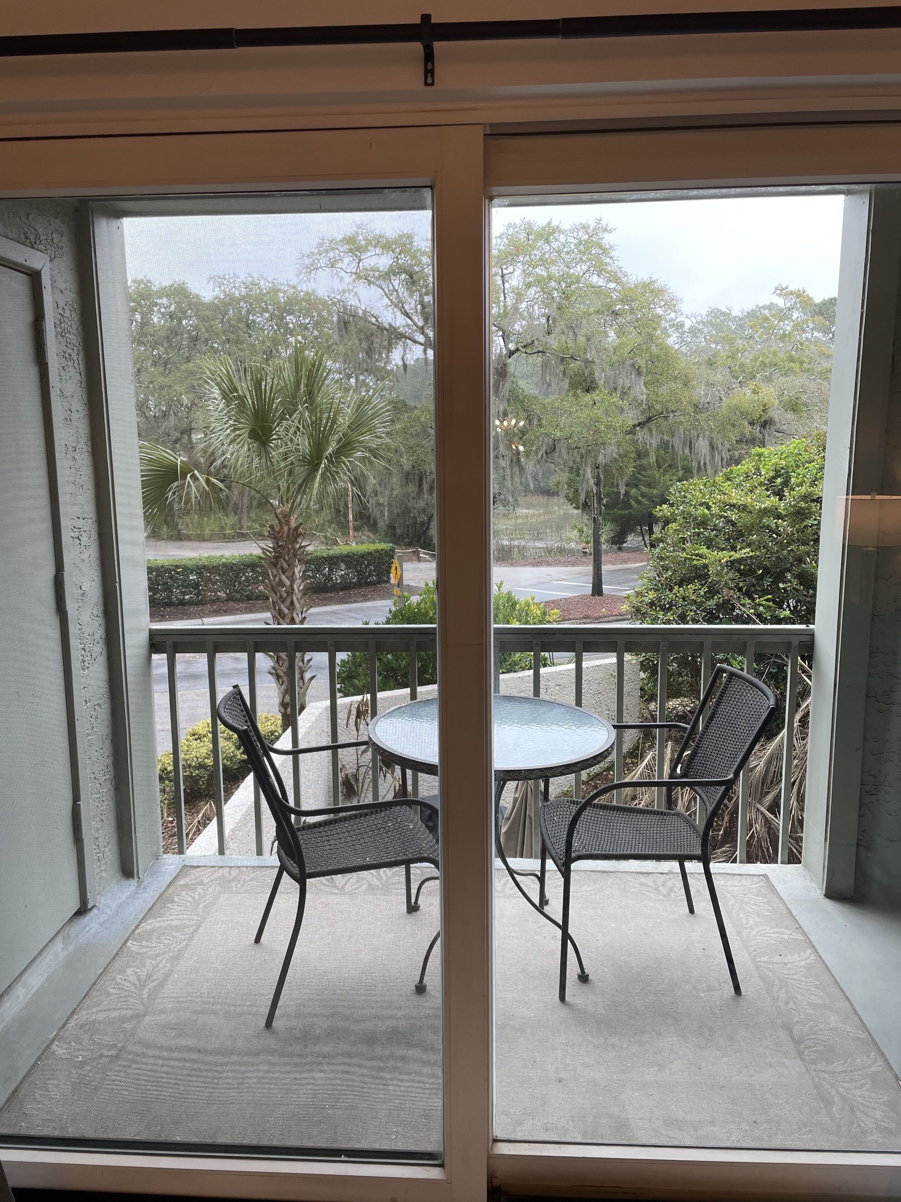 Uma varanda com mesa redonda e duas cadeiras, com vista para um jardim com árvores e uma estrada.